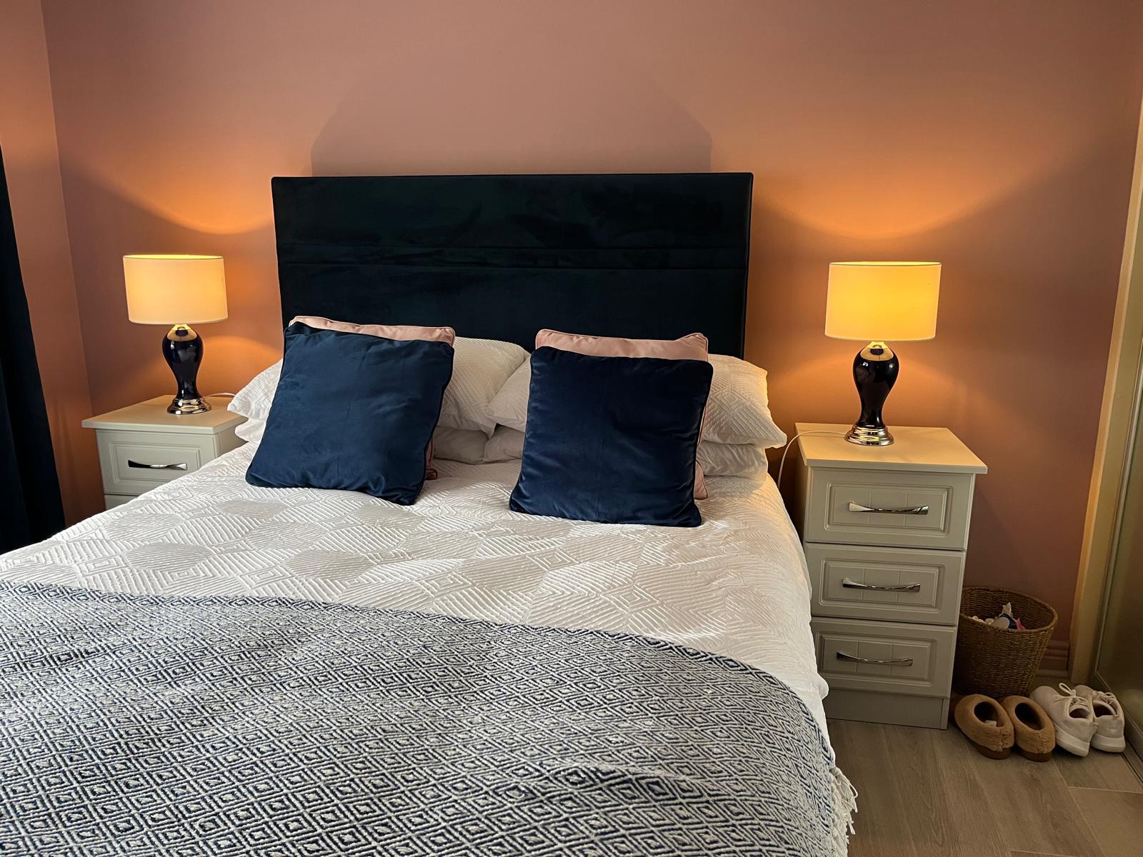 a bedroom with pink walls, a navy blue, velvet bed with two side tables. The side lamps have deep blue bases and white shades and are switched on. The bedclothes are white and there are two navy throw cushions.